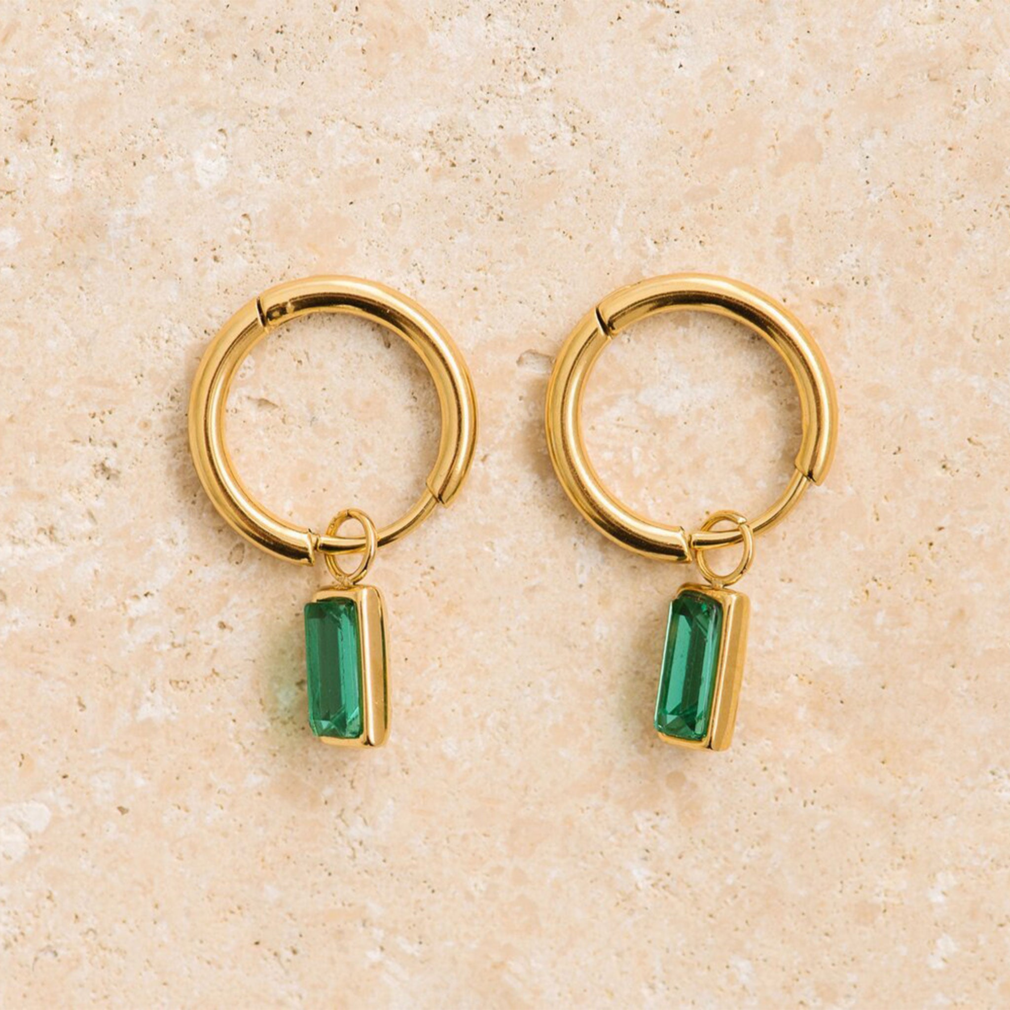 On a tan background is gold hoop earrings with an emerald cut emerald stone hanging from it