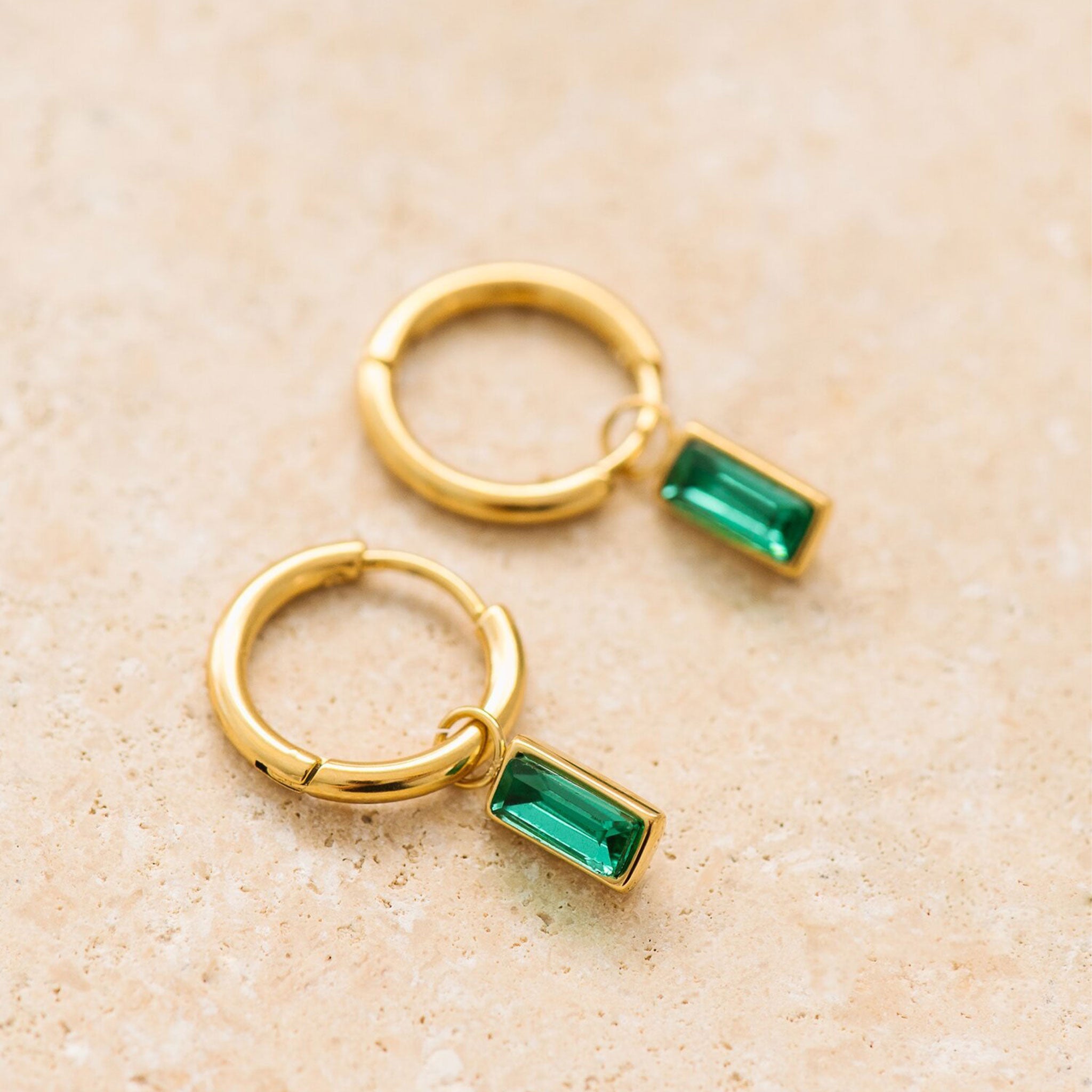 On a tan background is gold hoop earrings with an emerald cut emerald stone hanging from it. 