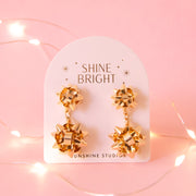 A pair of gold holiday bow shaped earrings that have a smaller gold bow and a dangling larger bow attached to the bottom. The packaging reads, "Shine Bright Sunshine Studios".