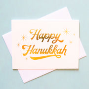 An ivory greeting card that reads happy Hanukkah in cursive gold foil and surrounded by six starbursts.