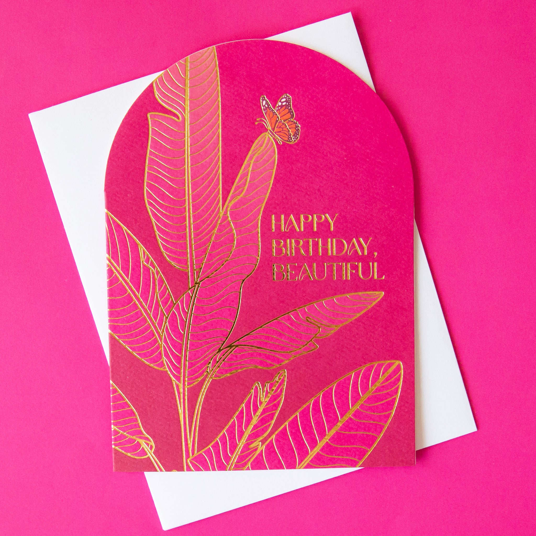 A bright pink arched shaped card with gold foiled illustrations of a banana leaf plant and gold text that reads, &quot;Happy Birthday Beautiful&quot;.