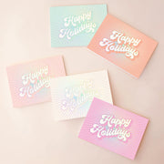 On a tan background is five greeting cards in different colors that read, "Happy Holidays" with holographic rays coming out from the text in the center. This pack of card includes one pink, white, light pink, peach and teal card. 