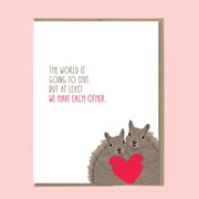 A white card with two squirrels at the bottom holding a heart and text that reads, 'The world is going to shit. but at least we have each other'. 