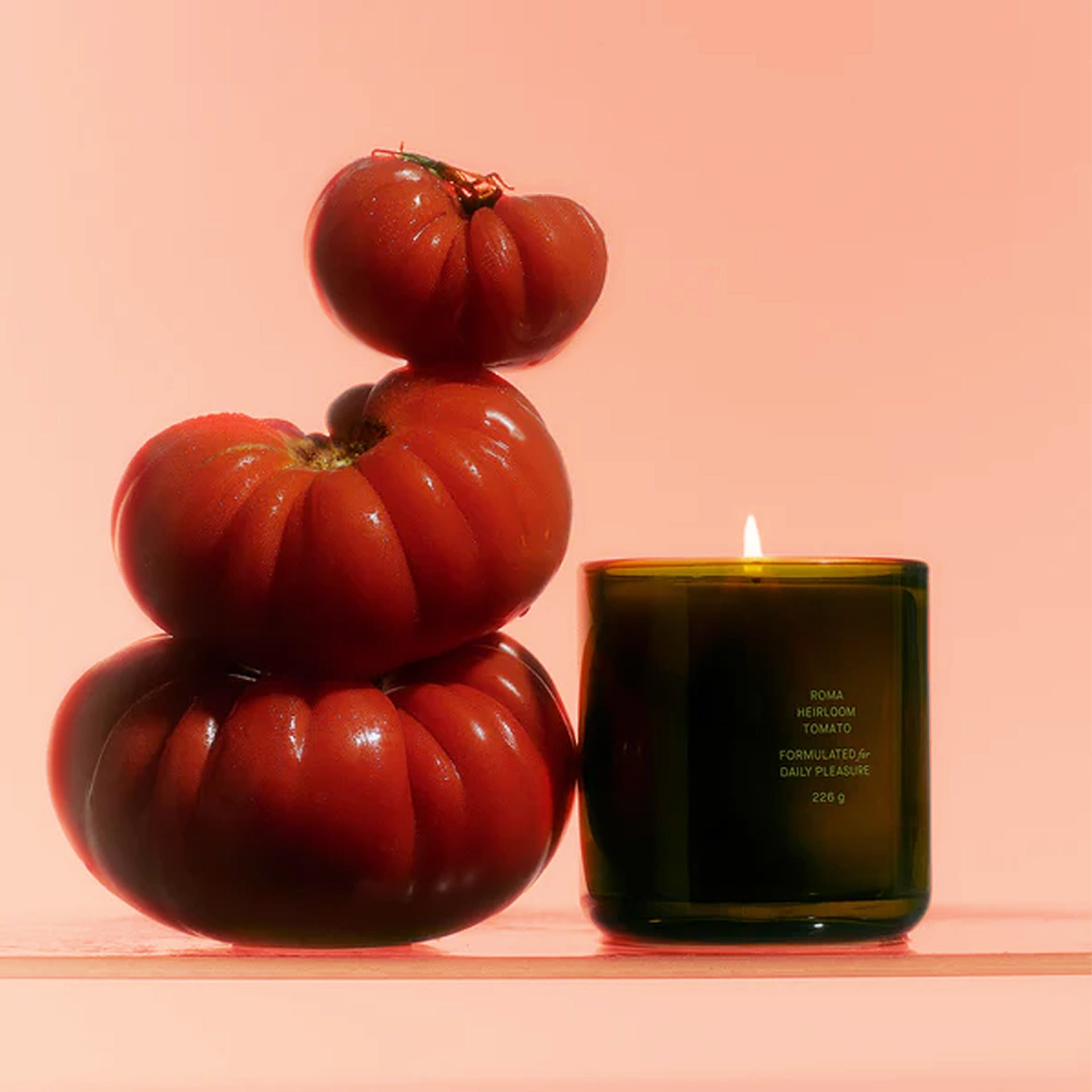 A green glass candle with small text in the front that reads, "Roma Heirloom Tomato Formulated for Daily Pleasure" surrounded by red tomatoes.