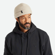 A knit ribbed beanie in a beige color with a folded brim and a black label on the front right left corner.