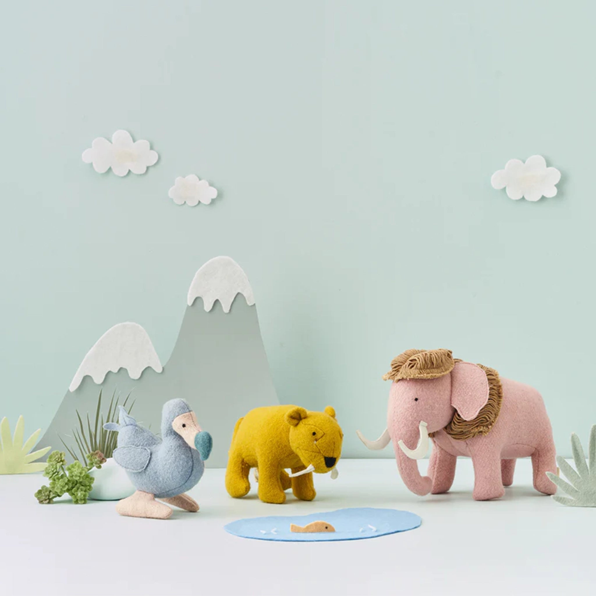 On a light blue background is a set of extinct stuffed animals. A blue dodo bird, a yellow sabre tooth tiger, and a pink mammoth. 