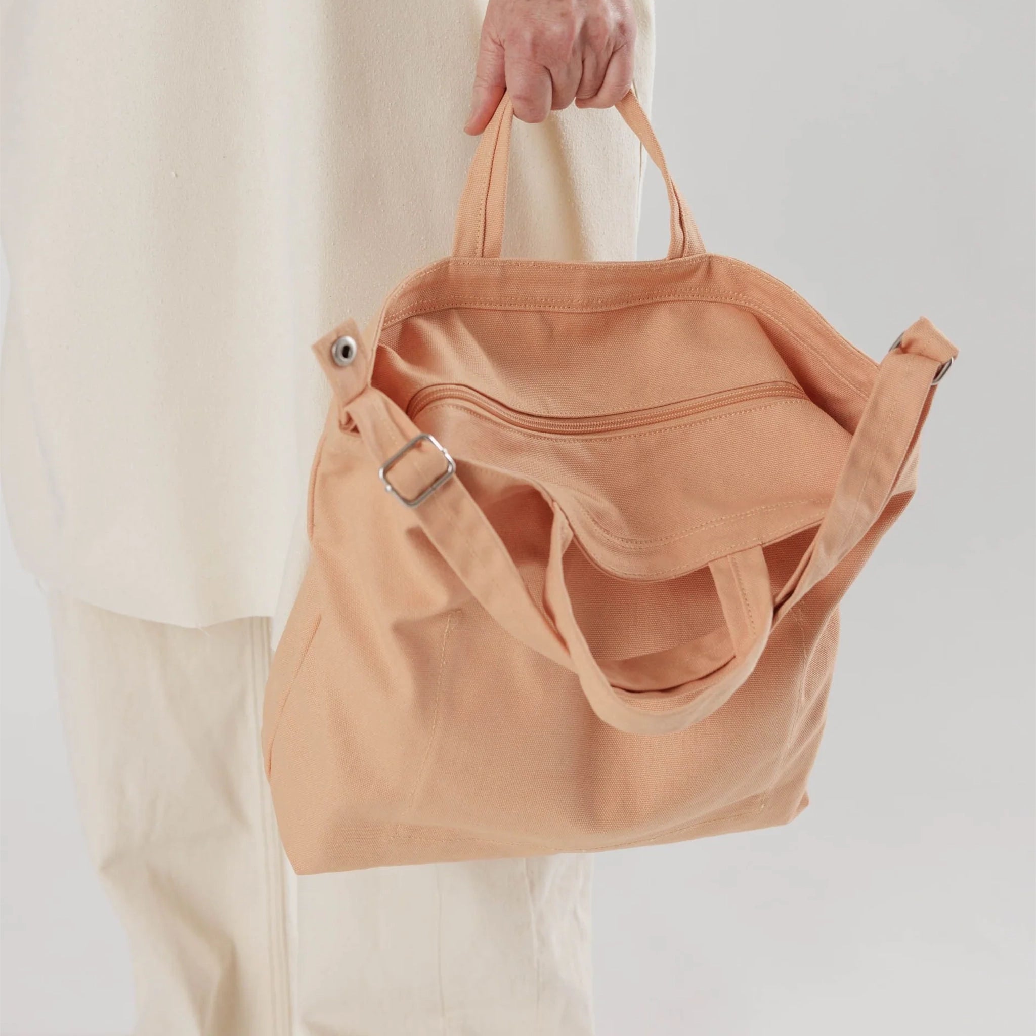 A square canvas tote bag with a zipper and two handles and a longer over the shoulder strap in a salmon color.