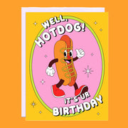 On an orange background is an orange and pink card with an illustration of a smiley face hot dog with text above and below that reads, "Well, Hotdog! It's Ur Birthday".