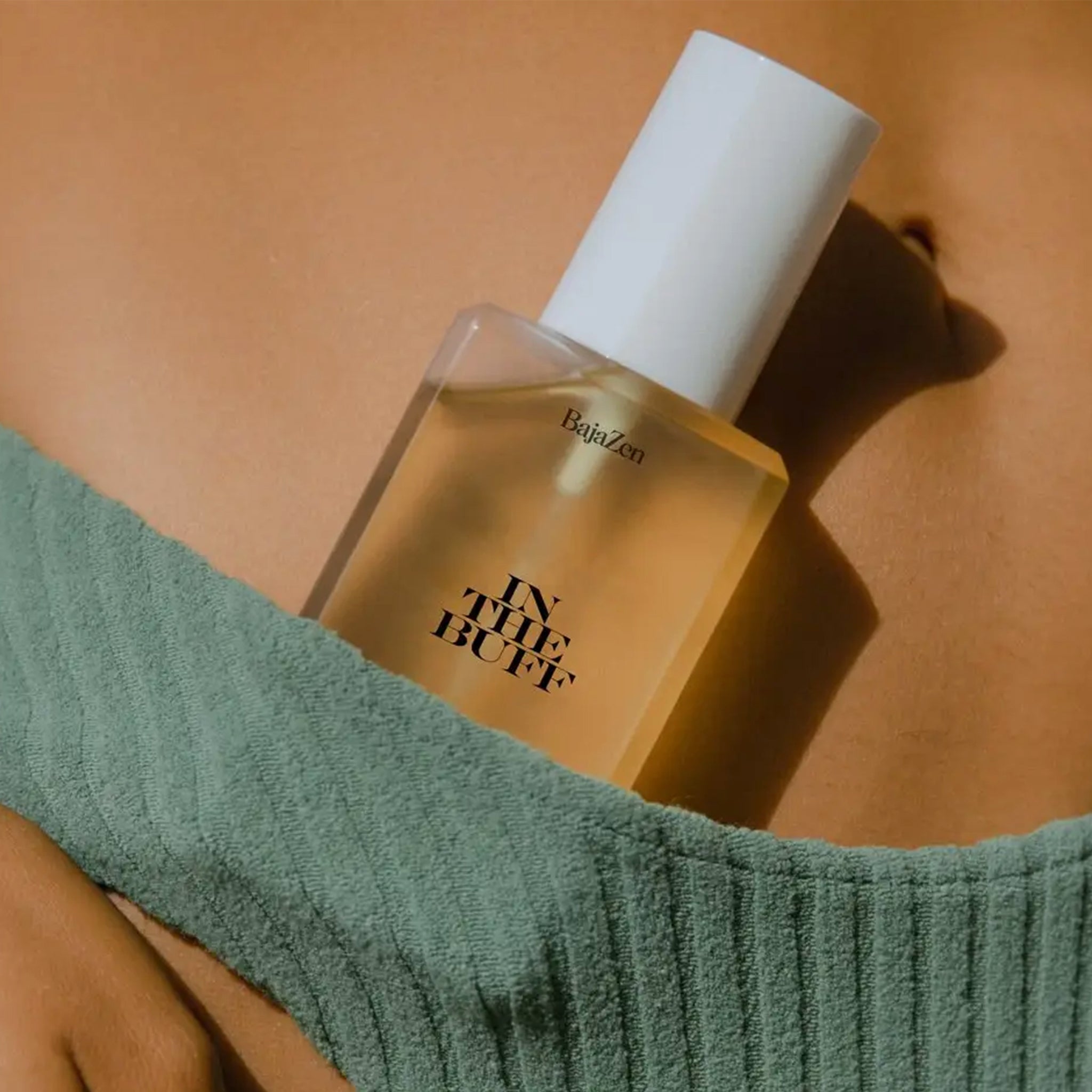 A clear packaging of body oil with a white lid and black text that reads, "In The Buff". 
