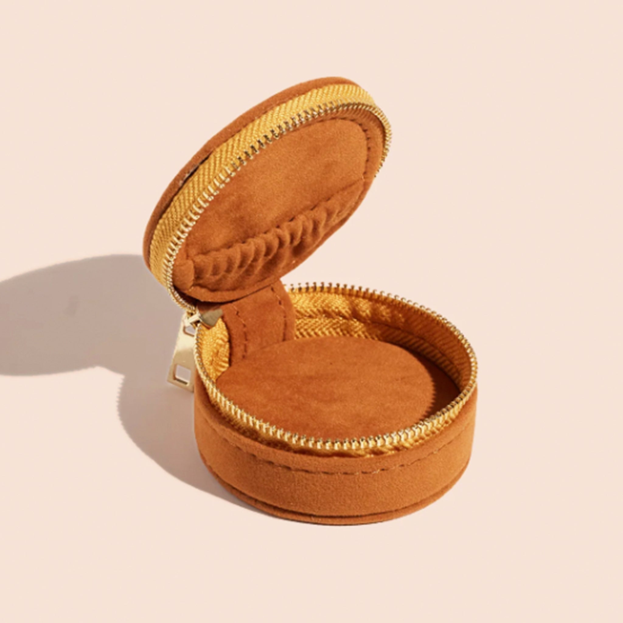 A circular suede burnt orange jewelry case with a gold zipper and a sun design on top.