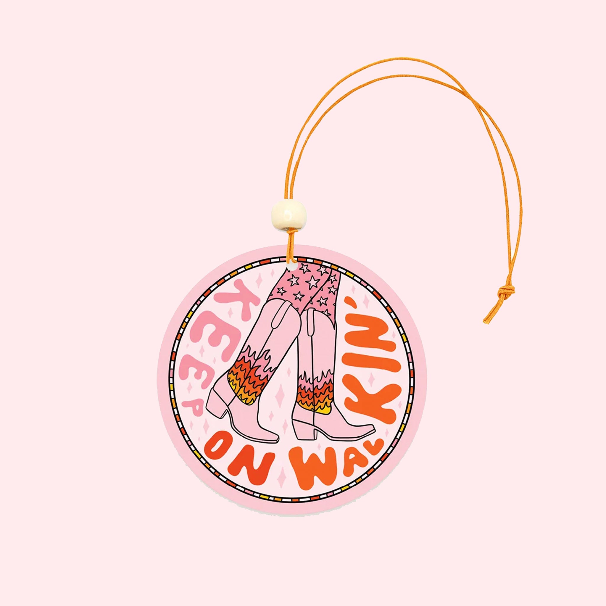 A light pink circle air freshener with a elastic loop for hanging and a cowgirl boot graphic with fire details on them and text that reads, "Keep On Walking'" in keep and red text around the edge of the air freshener.