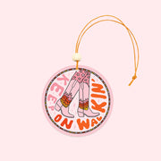 A light pink circle air freshener with a elastic loop for hanging and a cowgirl boot graphic with fire details on them and text that reads, "Keep On Walking'" in keep and red text around the edge of the air freshener.