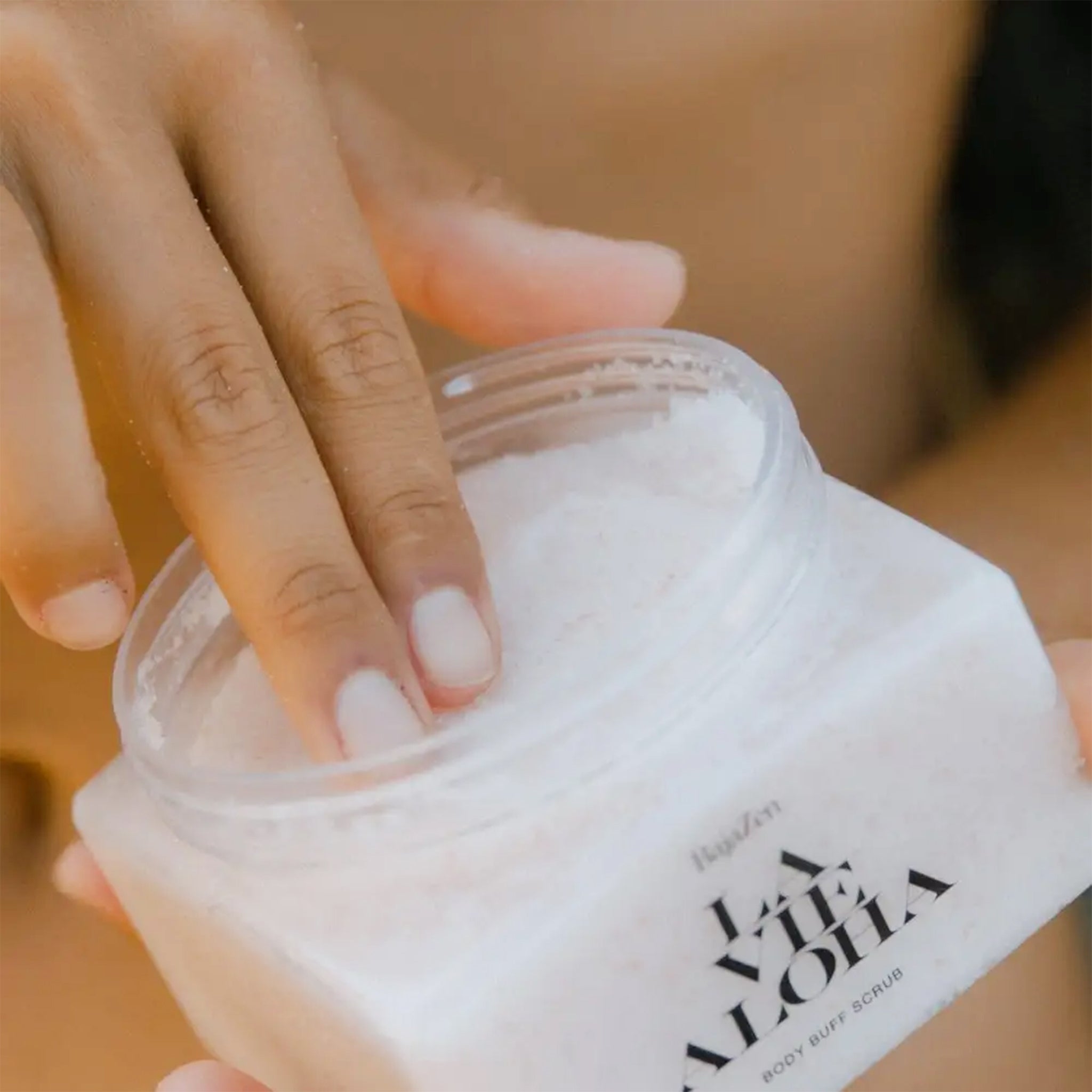 A clear container with a neutral body scrub inside, white lid and black text that reads, "La Vie Aloha".