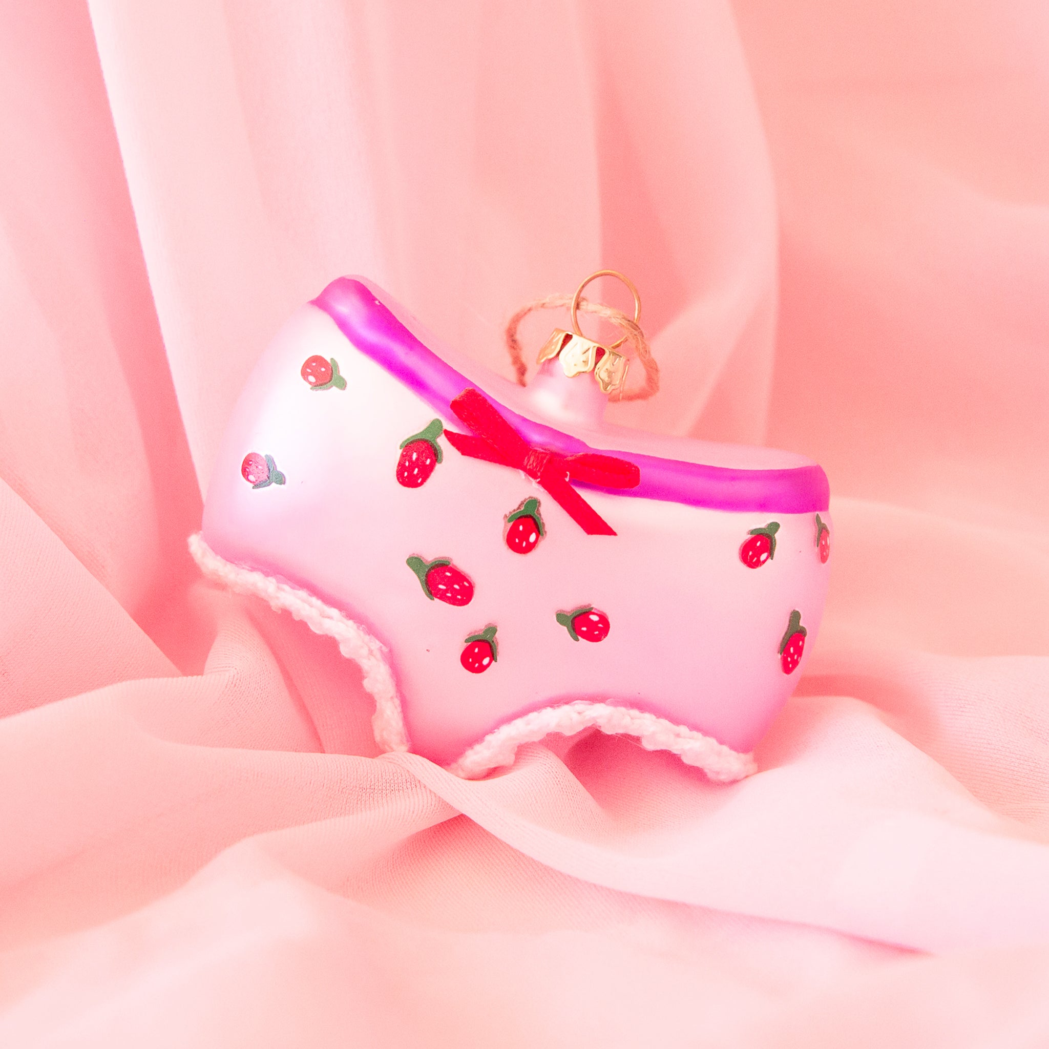 On a pink background is a glass ornament in the shape of underwear with pink details and a strawberry design. 