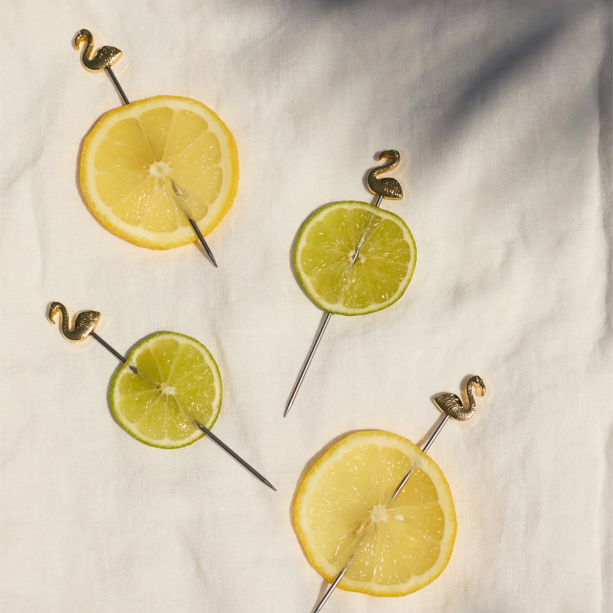 A set of four gold cocktail picks with flamingo shapes at the end speared through slices of lemons and limes. 