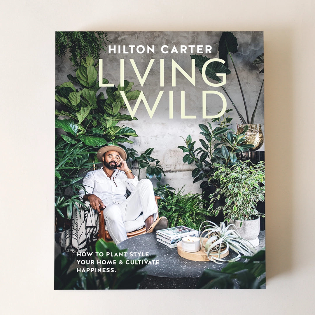 On a neutral background is a book cover with the title "Living Wild" along with greenery around the edges and the background and a photograph of the other of the book in the center.