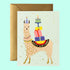 A light blue card with an illustration of a llama wearing a yellow happy birthday saddle and carrying a tower of multi colored wrapped gifts and a cake with a single candle on top. Also included is a coordinating envelope.