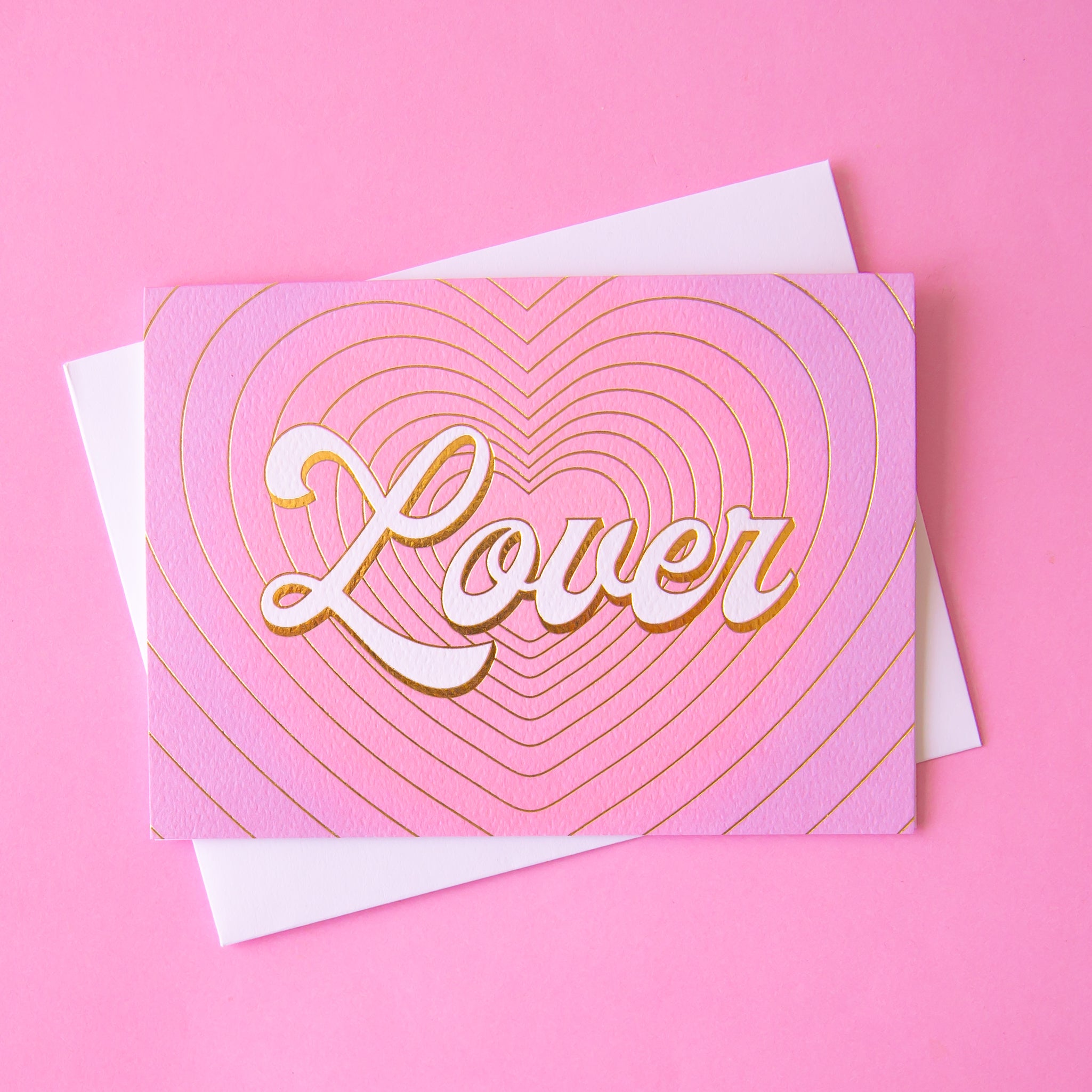 On a pink background is a pink and purple gradient card with gold foiled heart shaped outlines and ivory text that reads, "Lover". 