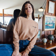 Woman with dark hair in a rust colored sweatshirt with "Mamacita" embroidered in black on the front, wearing jeans sitting near a mirror with vintage decor.