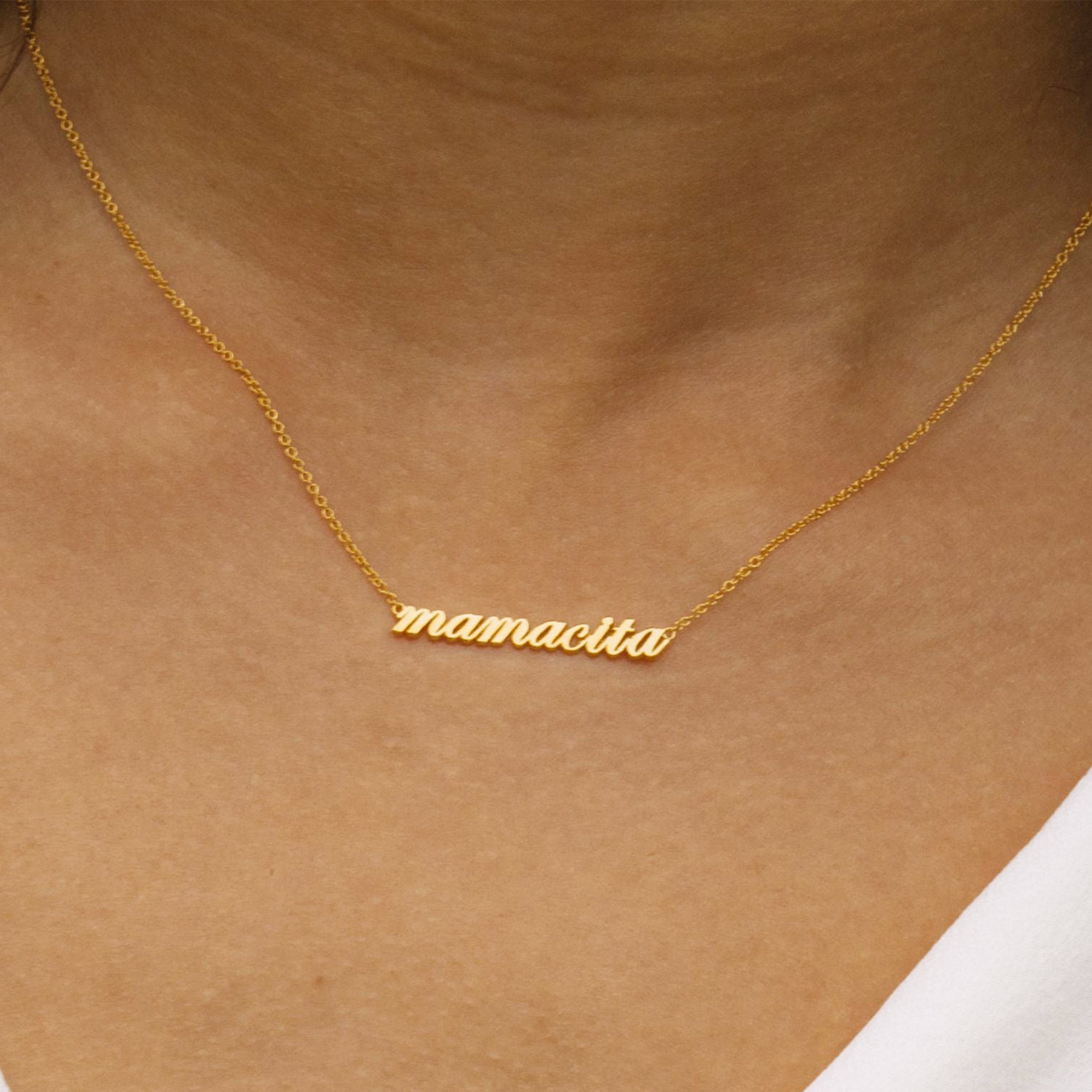 Zoomed in photo of a woman&#39;s neck. She is wearing a necklace that reads &quot;mamacita&quot; in lowercase script. A thin chain is connected on each side of the word.