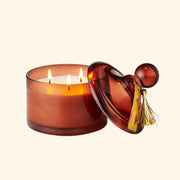 a brownish red glass jar candle with a lid and a festive gold tassel. 