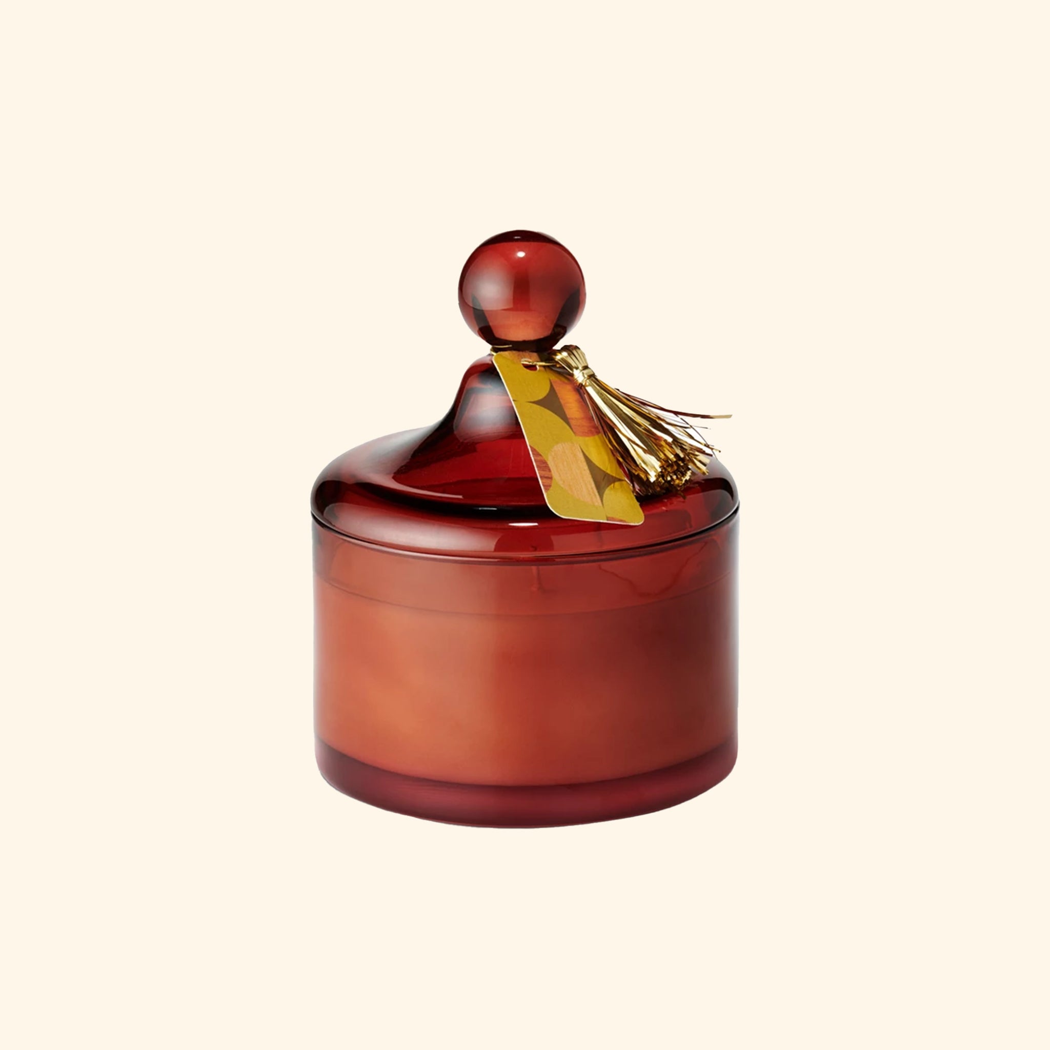 a brownish red glass jar candle with a lid and a festive gold tassel. 