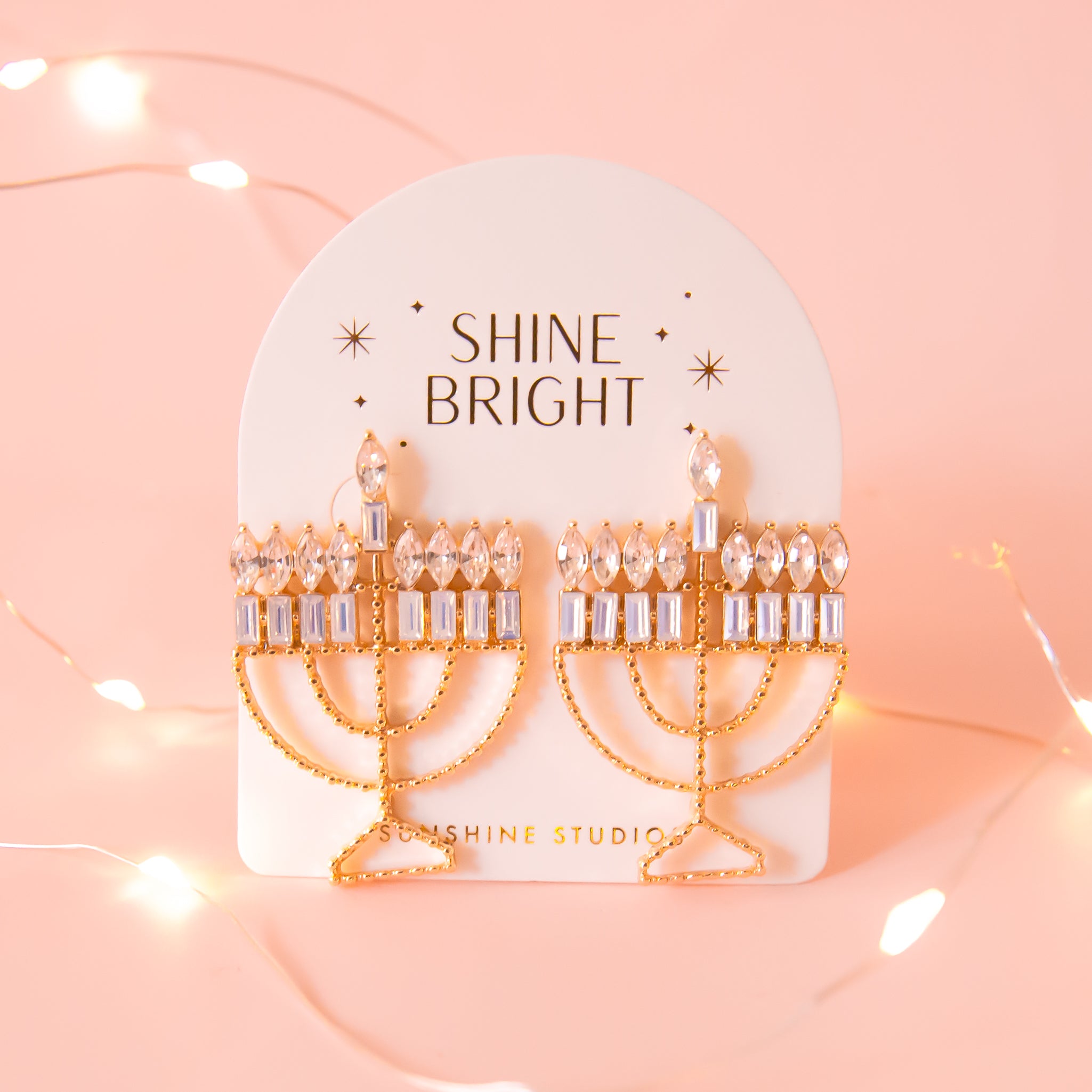 Menorah shaped gold earrings are attached to a cream colored card. the Card has gold foil stamp which reads Shine Bright and Sunshine Studios with glittering stars surrounding the text. The background of the photo is a soft warm pink color. 