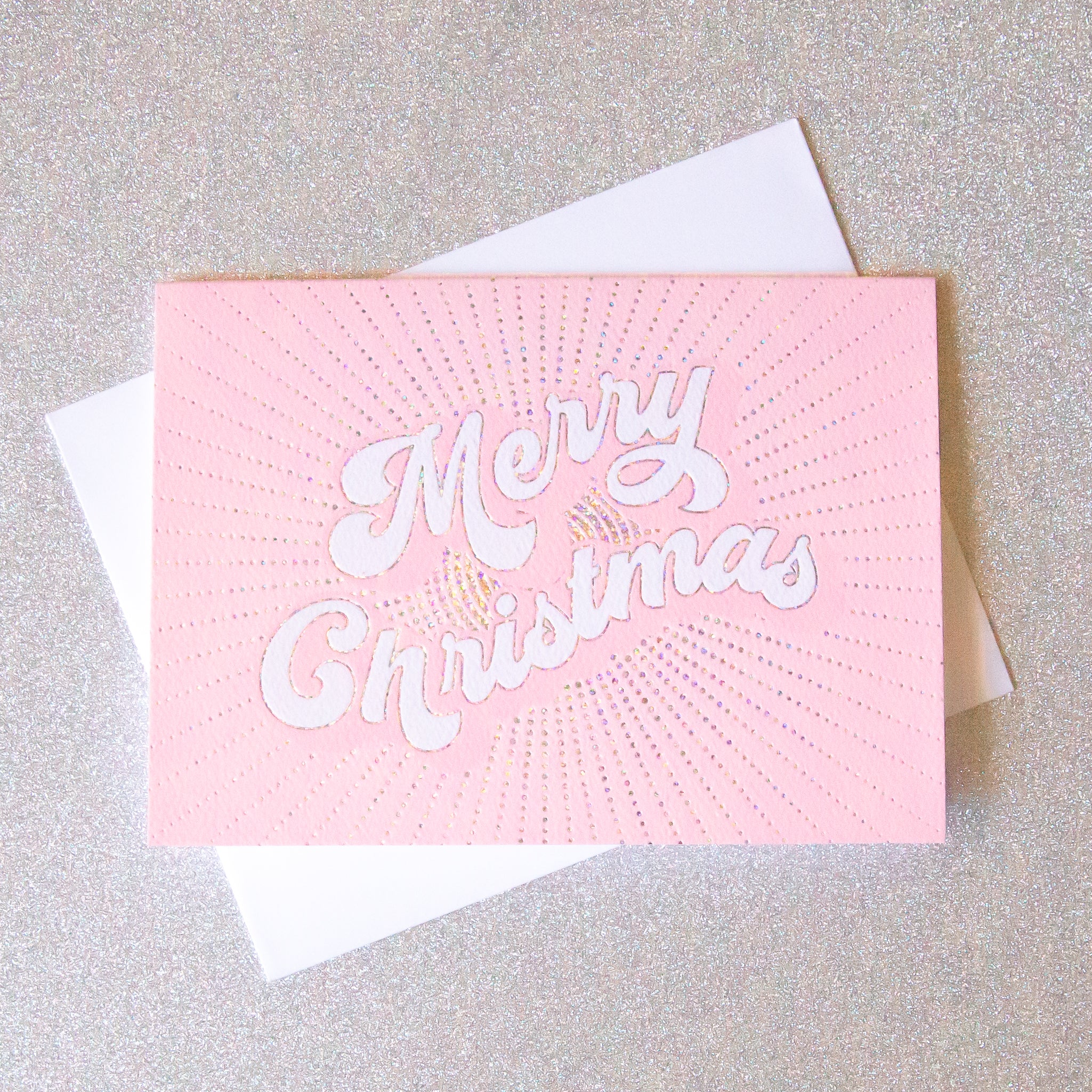 On a light pink background is a light pink greeting card with white text in the center that reads, "Merry Christmas" along with a white envelope.