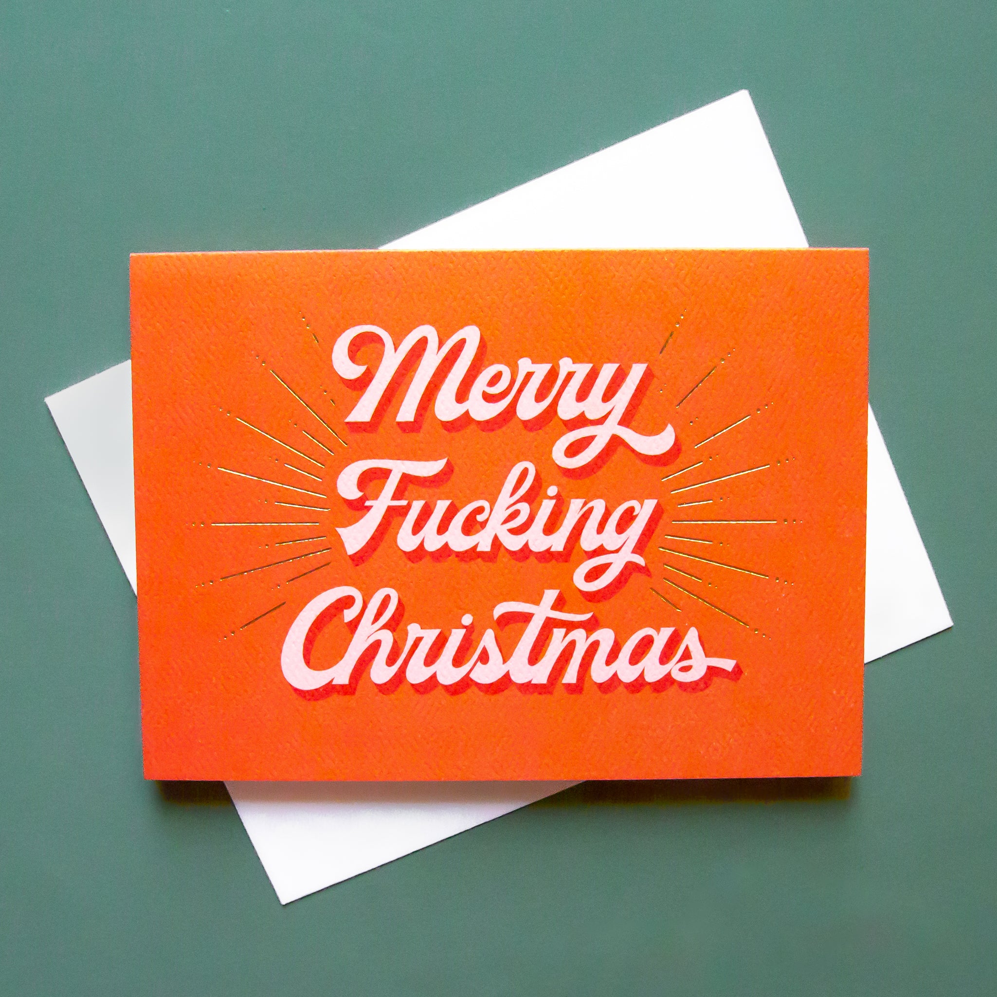 Red card that reads 'Merry Fucking Christmas' in soft pink lettering with dark red shadowing. A gold foil burst detailing beams from the text