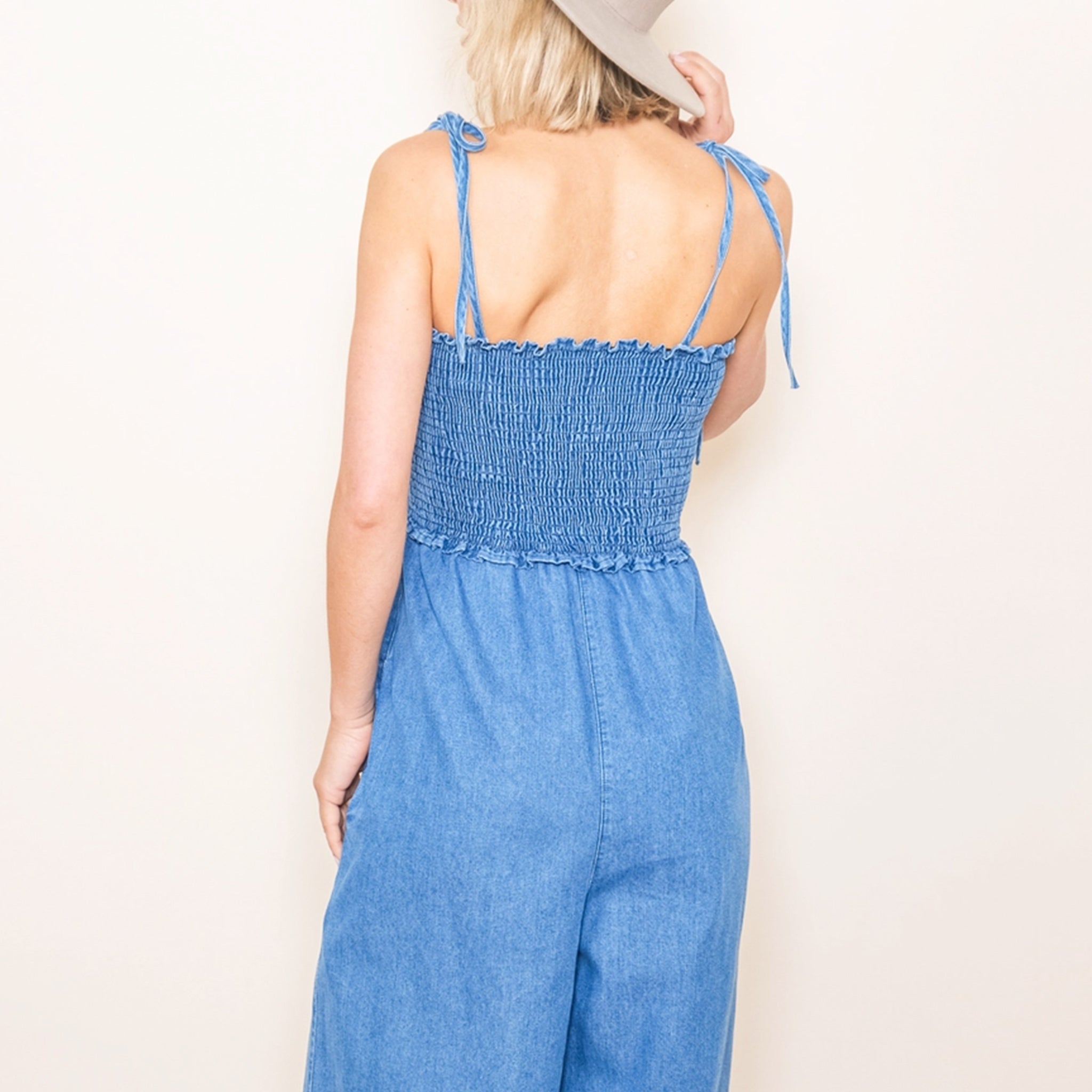 A mid blue shade of denim in a smocked jumpsuit with tie shoulder straps.