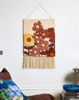 A hanging piece of woven wall art featuring rust and yellow colored floral prints and a fringe detail hanging from the bottom edge. 