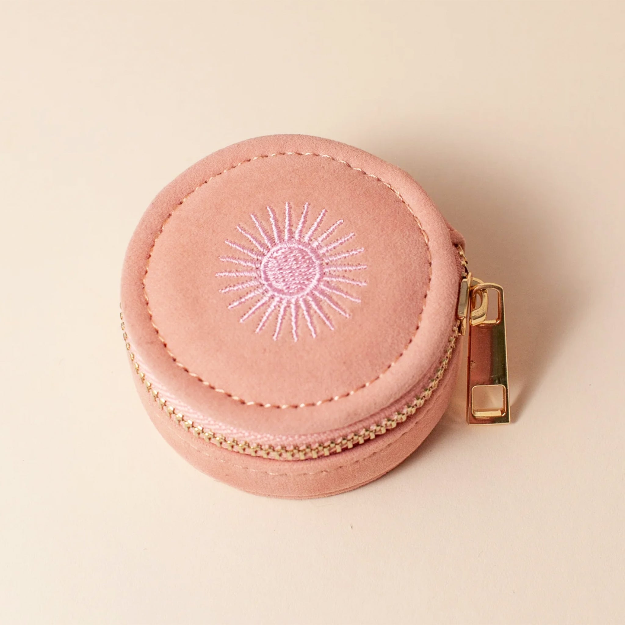 A rose colored circular jewelry case with a gold zipper and a sun design. 