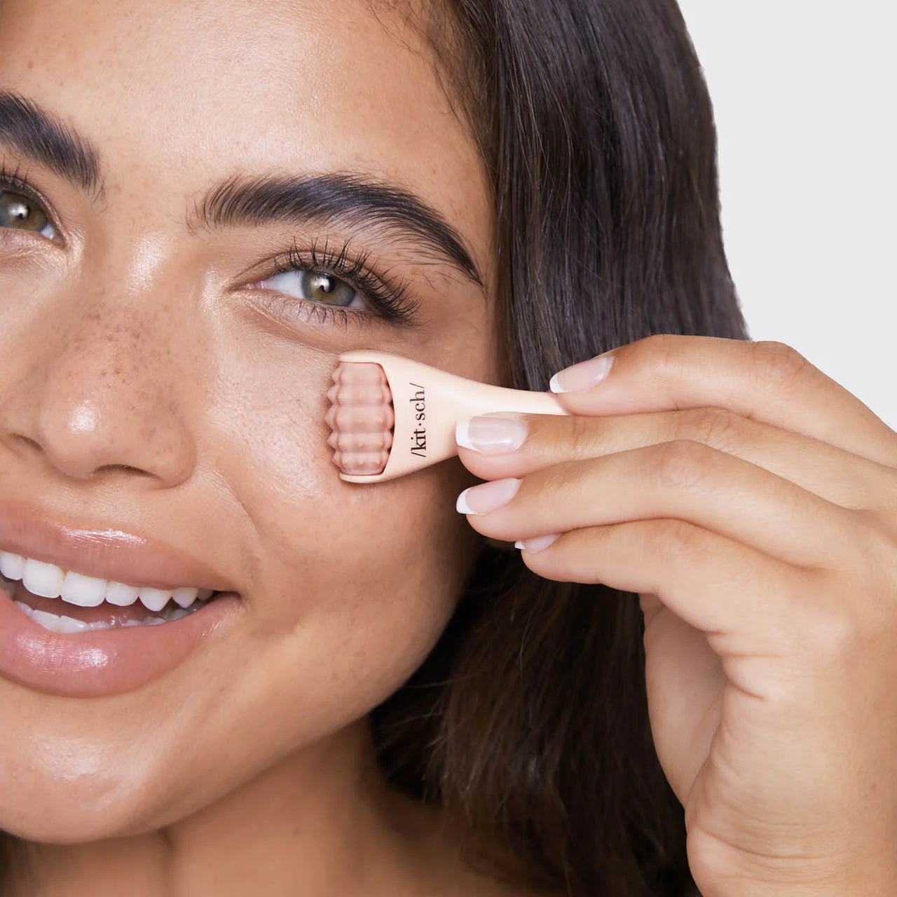 Woman with dark hair smiling holding a pink mini massage roller on cheek just below her eye.