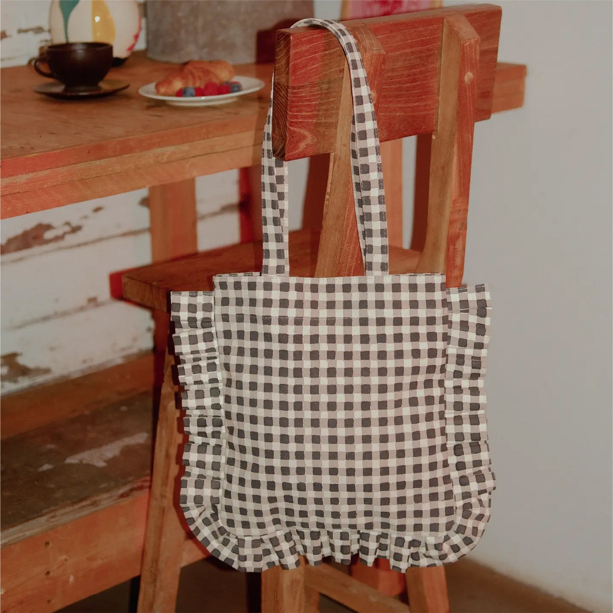 A black and white gingham print tote bag with ruffle details and shoulder straps. 