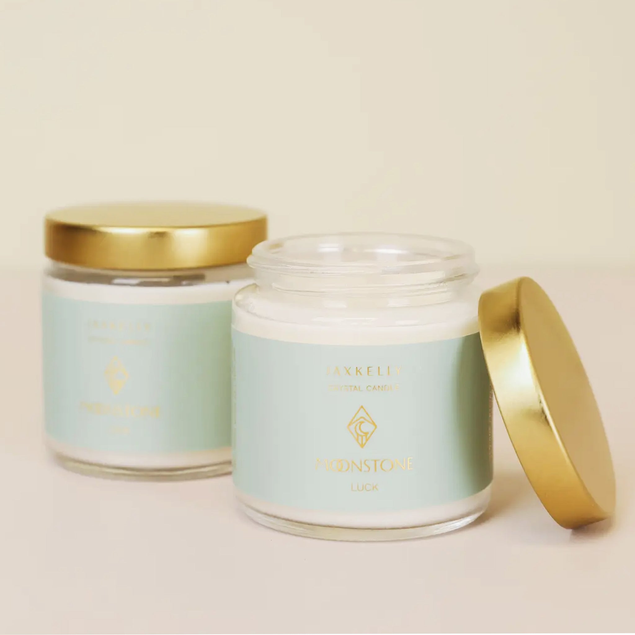 On a tan background is a clear glass jar candle with a light blue label and gold lid. 