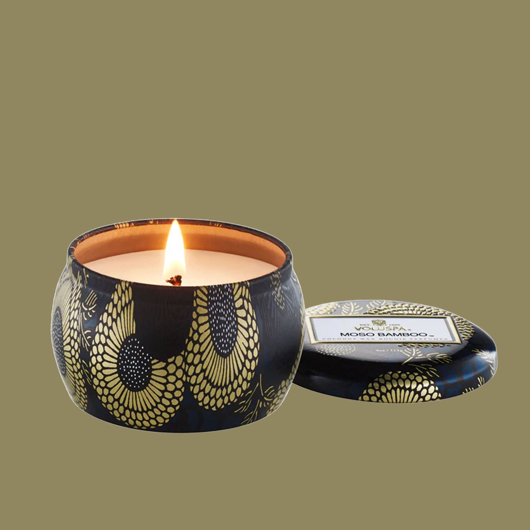 On a khaki background is a black and gold decorative tin single wick candle with a label on the lid that reads, "Voluspa Moso Bamboo".