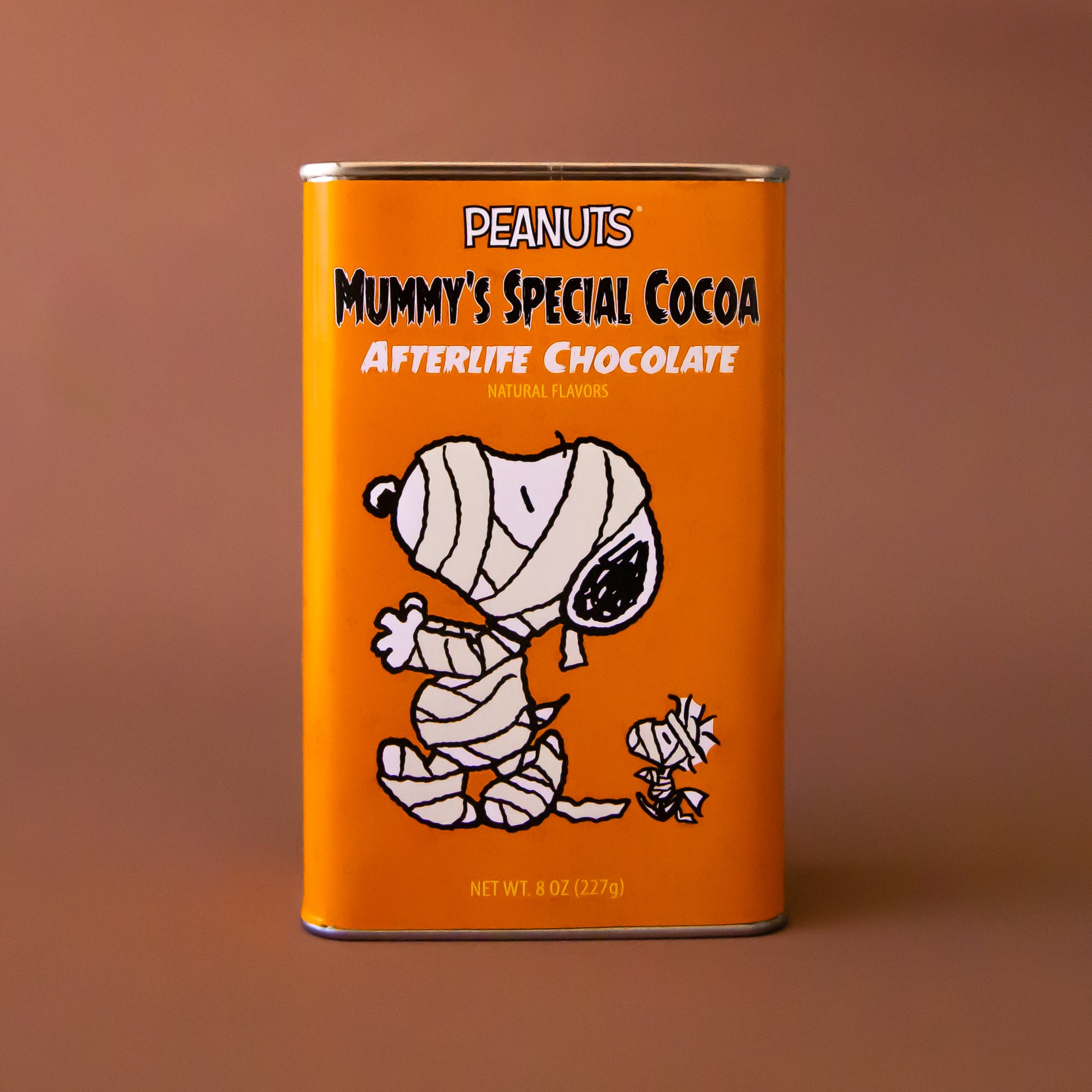 An orange rectangle tin of hot chocolate mix with a graphic of the Snoopy character wrapped up like a mummy and text that reads, 'Peanuts Mummy's Special Cocoa Afterlife Chocolate'. 