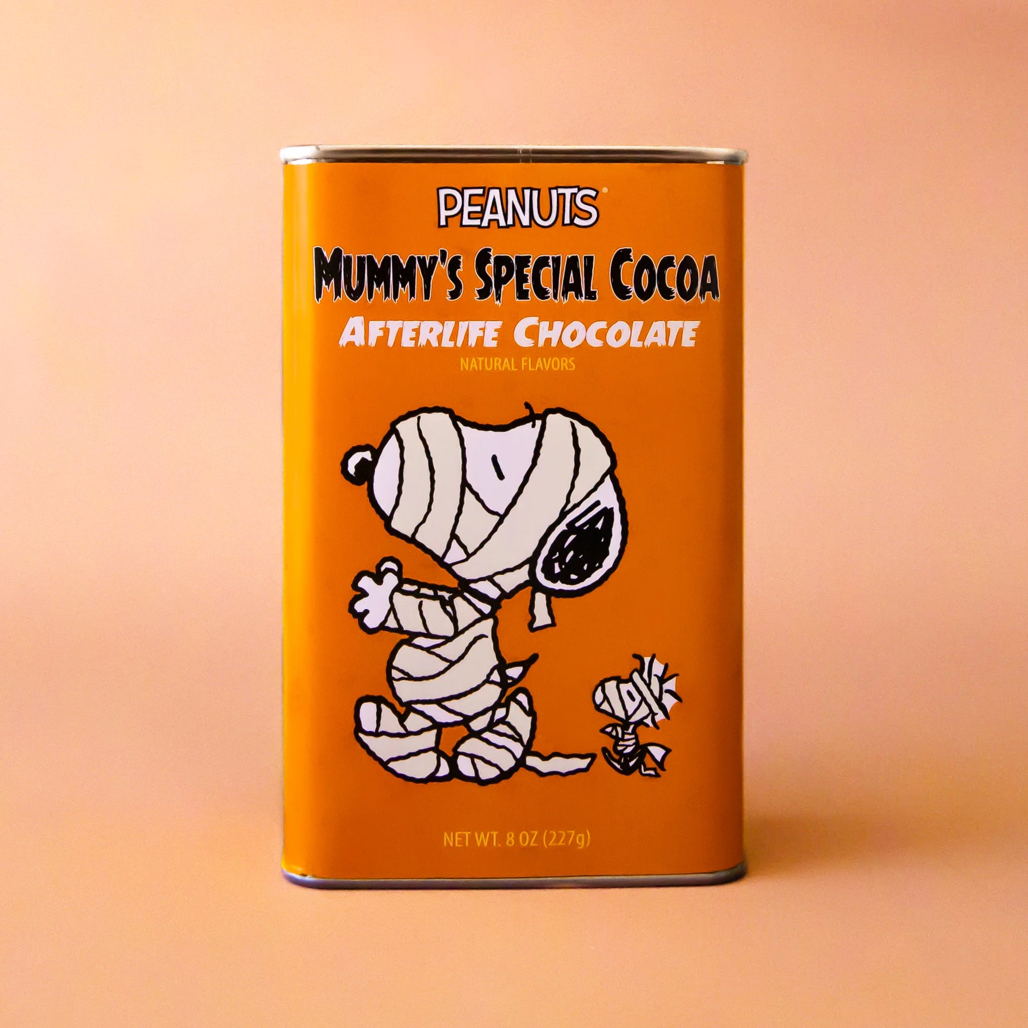 An orange rectangle tin of hot chocolate mix with a graphic of the Snoopy character wrapped up like a mummy and text that reads, &#39;Peanuts Mummy&#39;s Special Cocoa Afterlife Chocolate&#39;. 