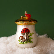 On a green background is an ivory and red glass ornament in the shape of a cookie jar with a mushroom design on it.