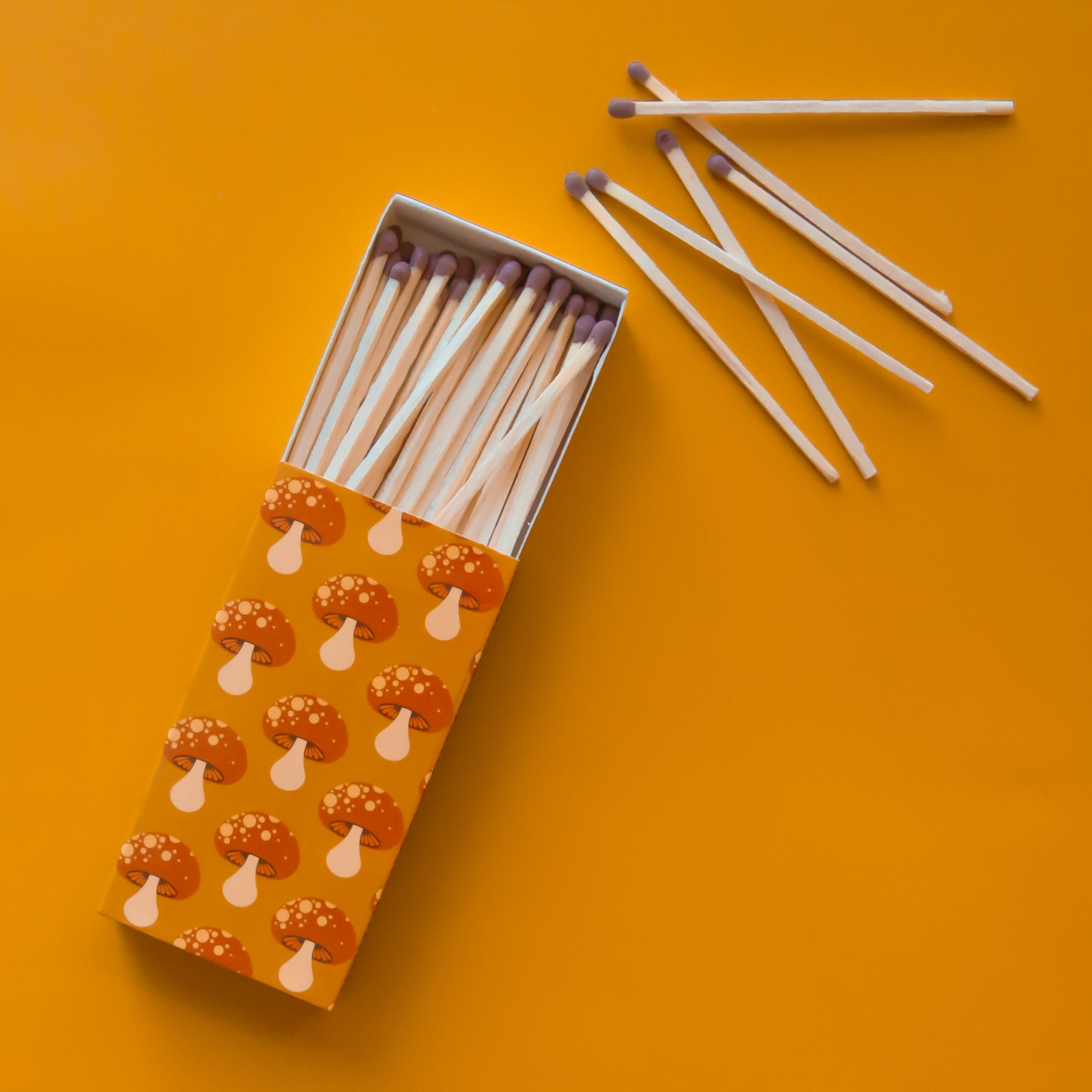 A rectangle box of matches with a repeating red mushroom design along all edges besides the top and bottom. The matches that are inside are also photographed here. They are wooden matches the same length as the box with a red striking tip for lighting. 