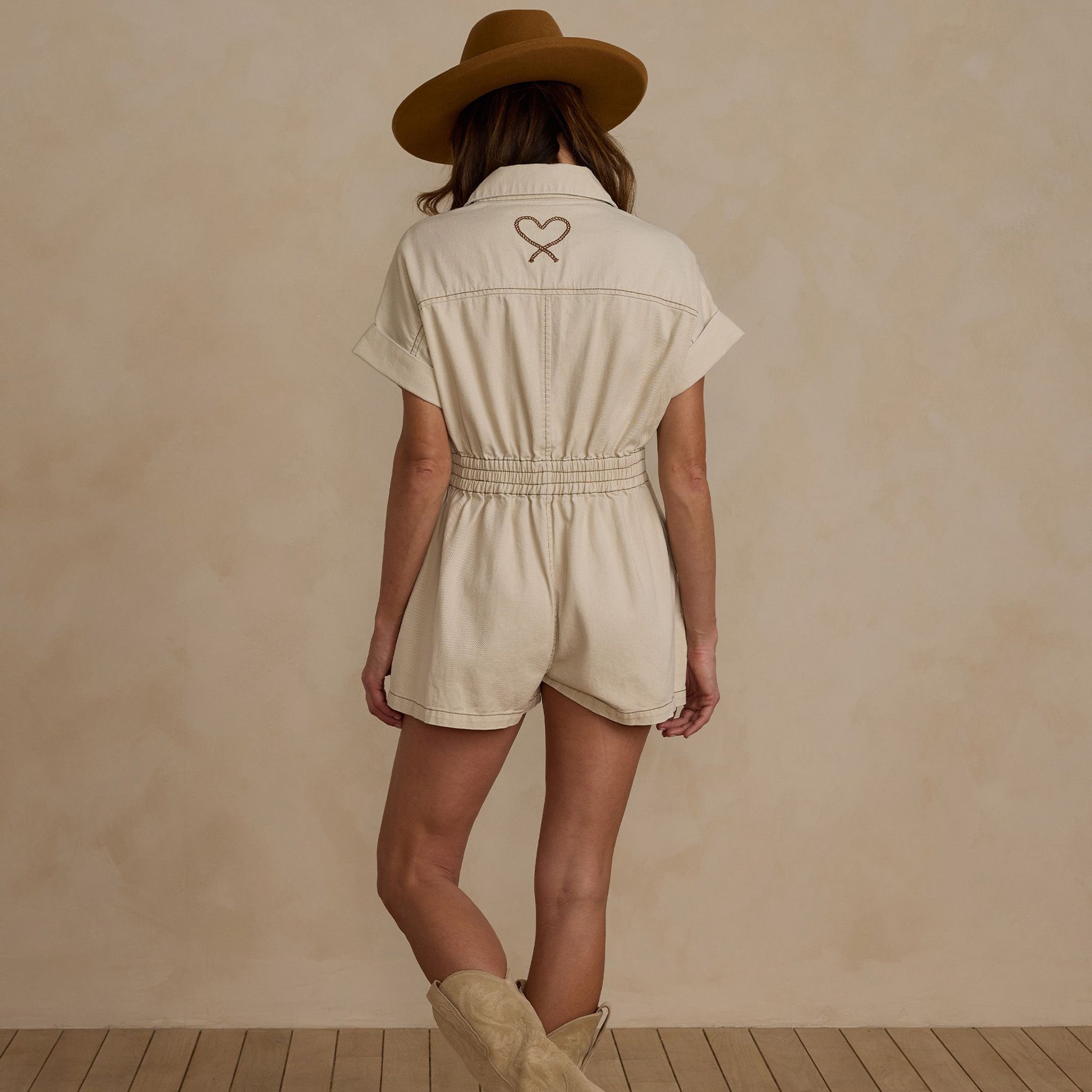 A natural colored short sleeve jumpsuit with a cinched waist and front pockets as well as a stitched rope heart detail on the back.