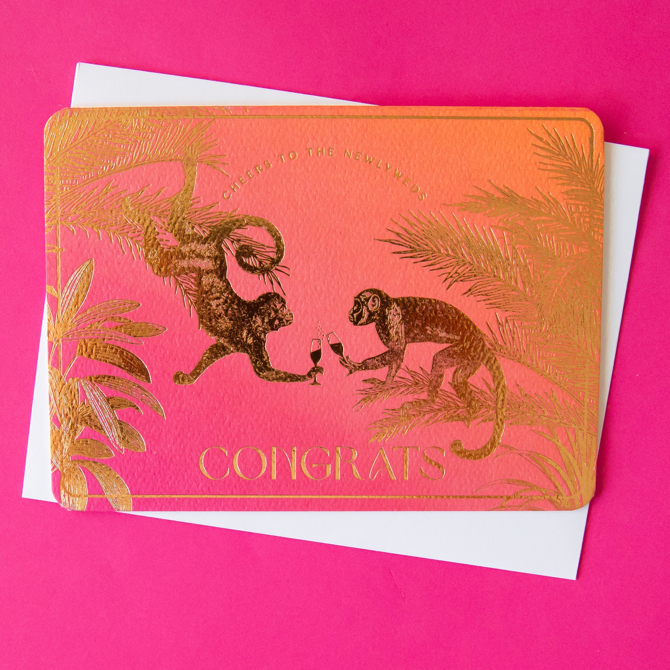A pink and orange ombre card with gold foiled text and design of monkey's in jungle foliage cheersing champagne flutes and text that reads, "Cheers to the Newlyweds Congrats". 