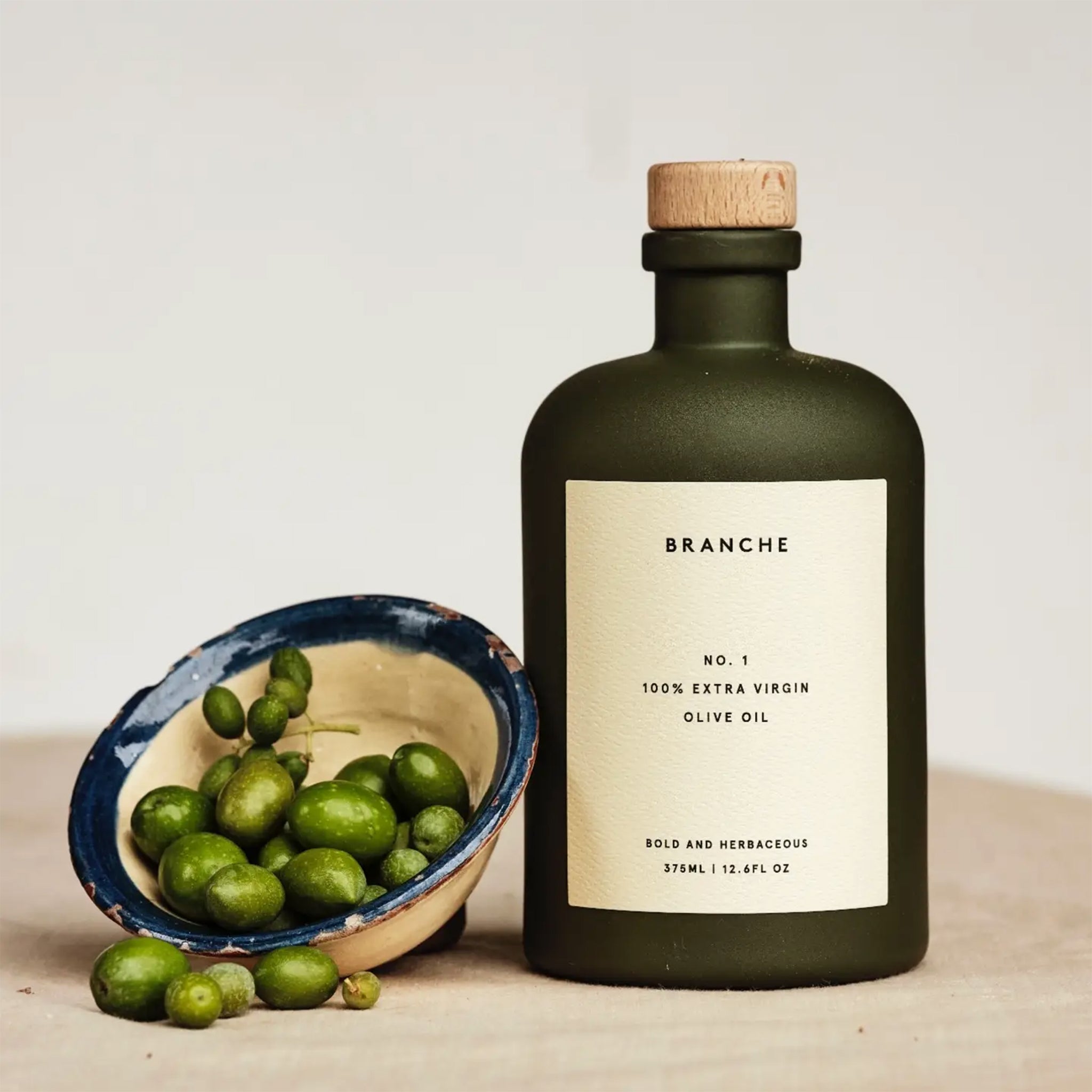 A green bottle of olive oil with a cork lid and an off white label with black text that reads, 'Branche No. 1 100% Extra Virgin Olive Oil'. 