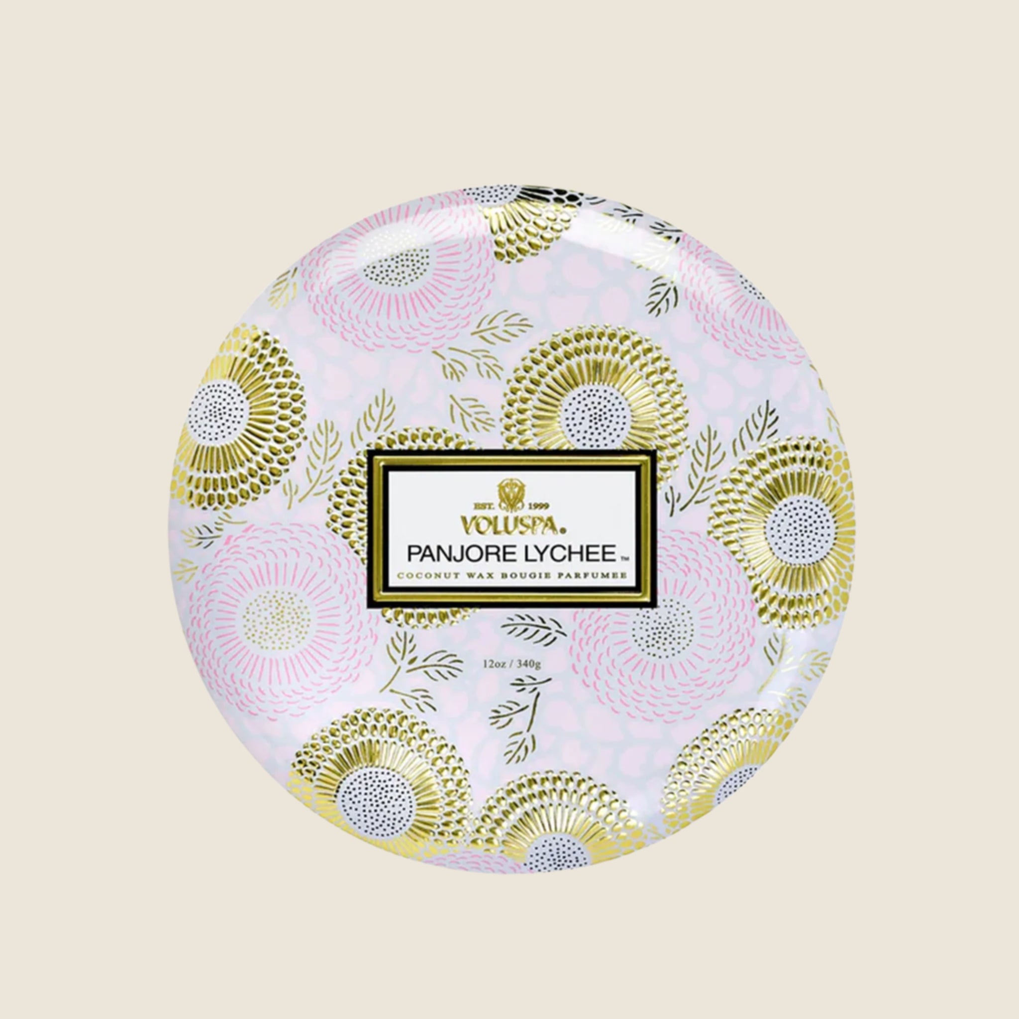 Inside the tin is a white candle with three white wicks. Leaning against the right side is a round tin lid. The lid has a light pink and gold floral pattern on it. In the middle is a white sticker with gold and black text inside.