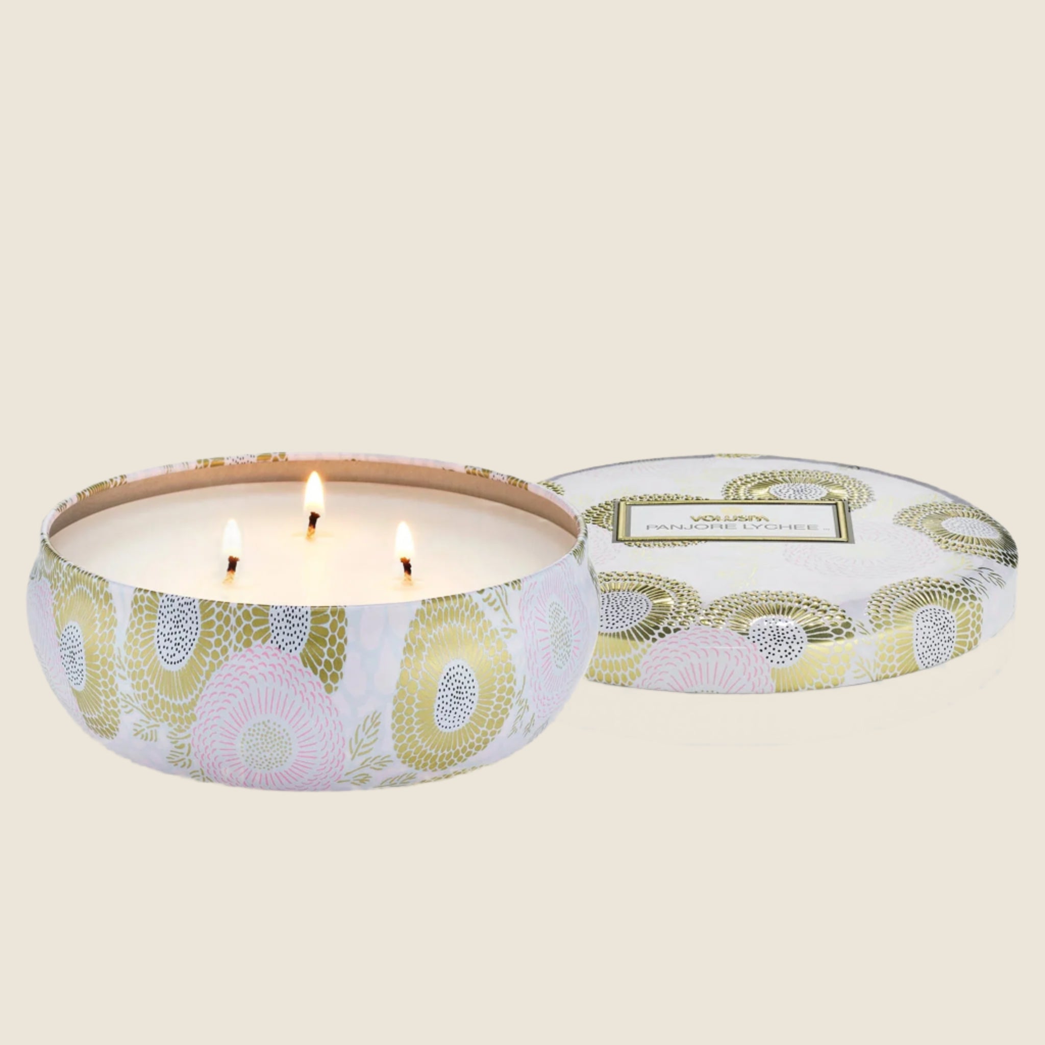 Inside the tin is a white candle with three white wicks. Leaning against the right side is a round tin lid. The lid has a light pink and gold floral pattern on it. In the middle is a white sticker with gold and black text inside.