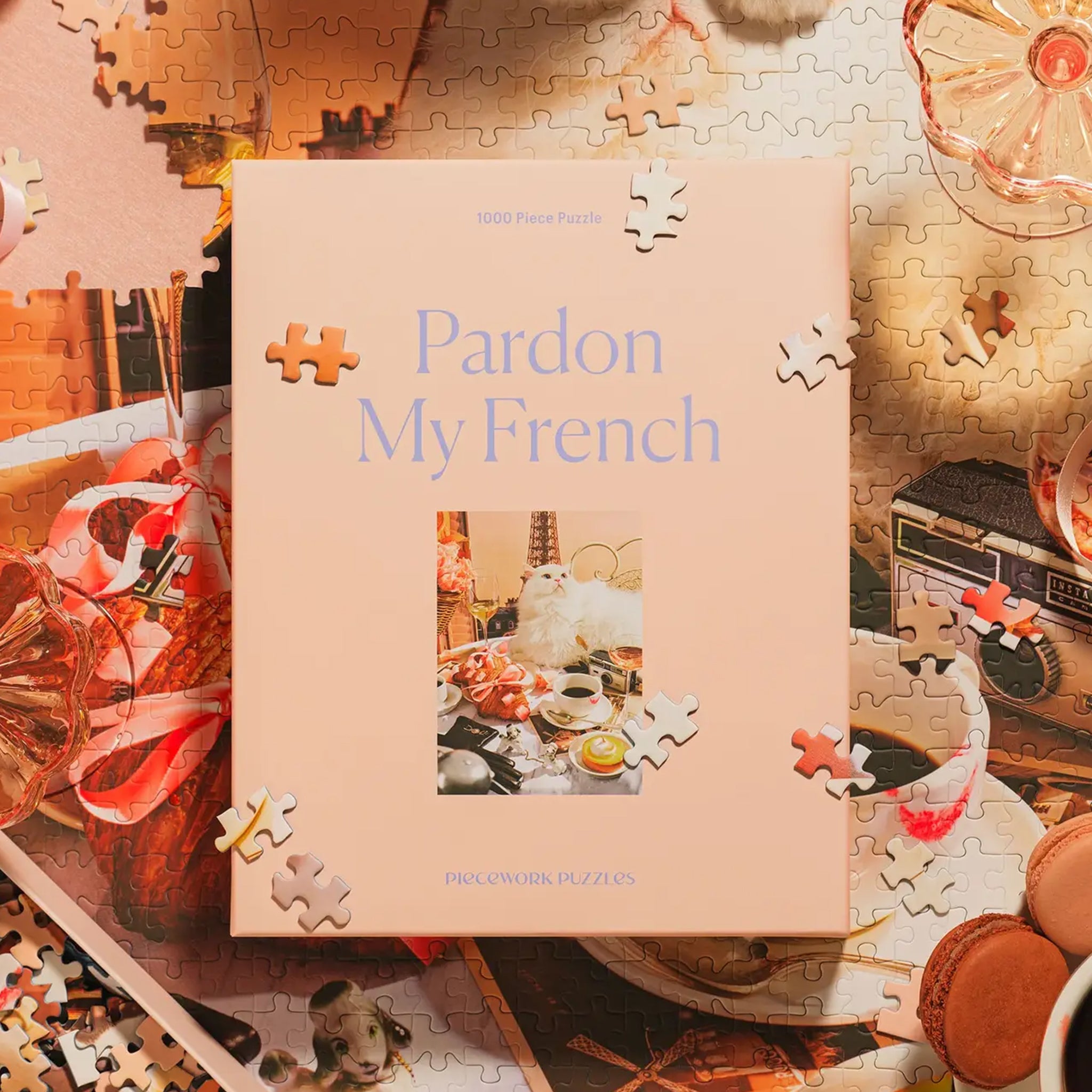 A peachy tan colored puzzle box with a 1000 piece puzzle inside with an image of a white fluffy cat surrounded by French goods such as croissants, macarons, champagne and the Eiffel Tower. 