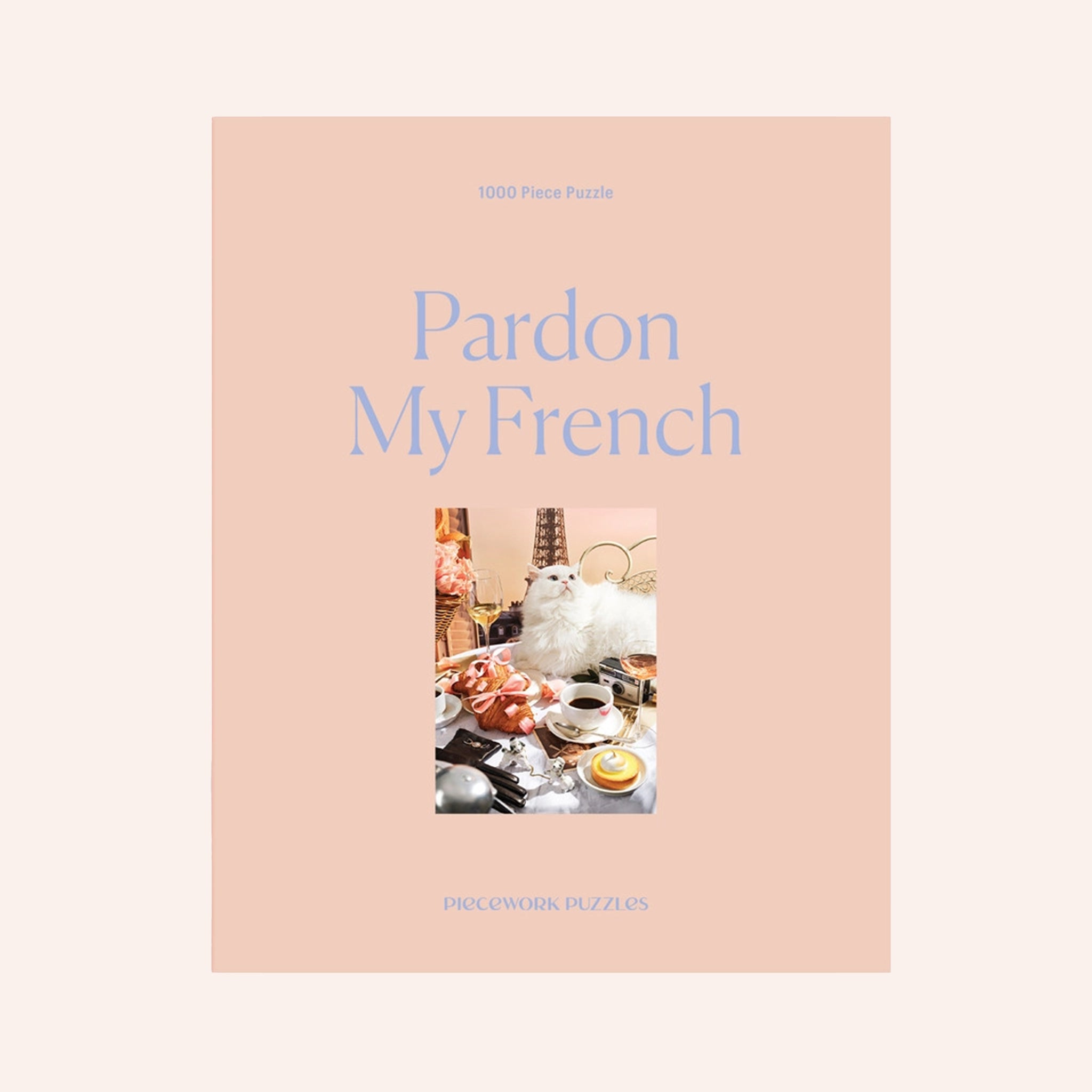 A peachy tan colored puzzle box with a 1000 piece puzzle inside with an image of a white fluffy cat surrounded by French goods such as croissants, macarons, champagne and the Eiffel Tower. 