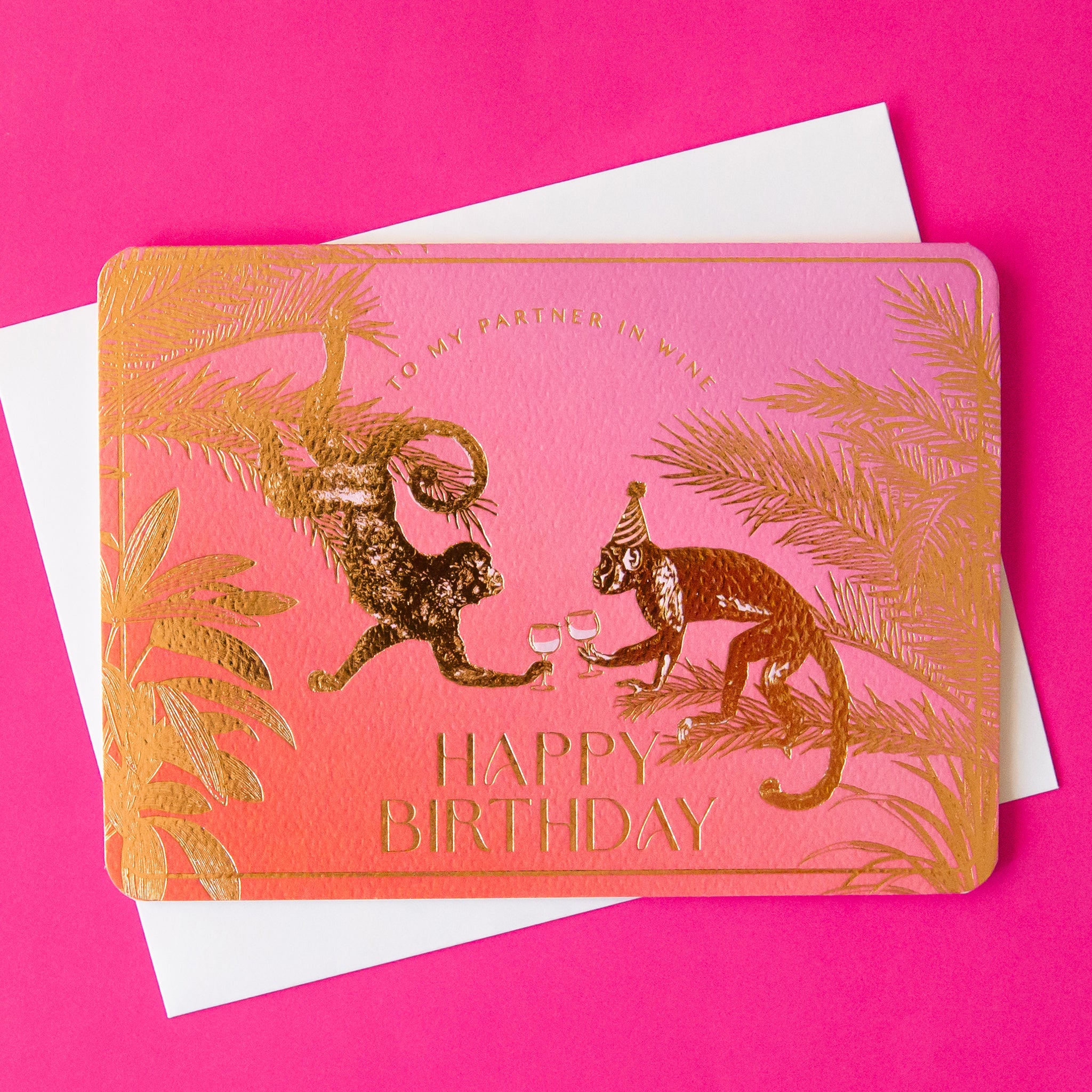 On a hot pink background is a pink and orange greeting card with gold foiled designs of monkeys cheersing and text that reads, &quot;To My Partner In Wine Happy Birthday&quot;.