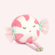 A darling peppermint candy shaped stuffed animal sits on a soft pink background.  She has little mint bows on either side of her face with pink fluff coming out the sides.  Her legs are sticking out and they are white and pink striped with little gold feet.  Her eyes are closed with long stitched black eyelashes and pink lips.