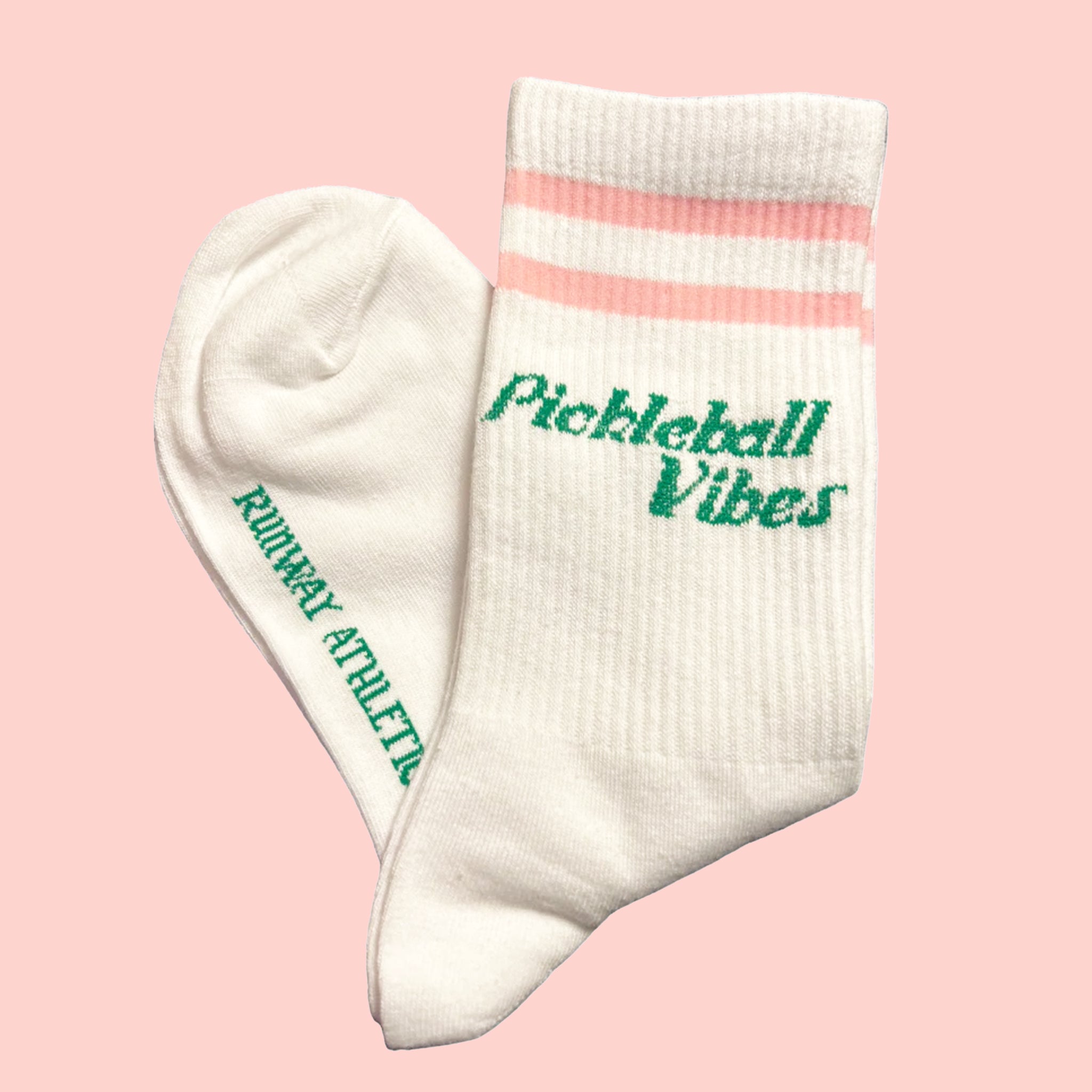 On a light pink background is a pair of white socks with pink stripes and green text that reads, &quot;Pickeball Vibes&quot;. 